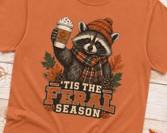 May include: An orange t-shirt featuring a graphic of a raccoon wearing a knit hat and a plaid scarf, holding a cup labeled Pumpkin Spice Latte. Below the raccoon, the text reads 'Tis the Feral Season' in bold, orange, and white block lettering.