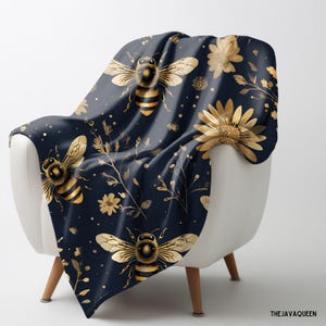 Pode incluir: Uma manta de arremesso azul marinho com um padrão de abelhas e flores douradas. A manta está drapeada sobre uma cadeira branca.
