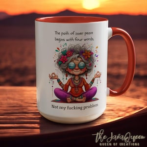 Könnte beinhalten: Ein weißer Keramikbecher mit leuchtend orangefarbenem Innenraum und Henkel. Die Vorderseite zeigt eine bunte Illustration einer Person mit grauem Haar und Brille, die in Lotusposition meditiert und von Blumen umgeben ist. Der Text lautet: The path of inner peace begins with four words, Not my fucking problem.