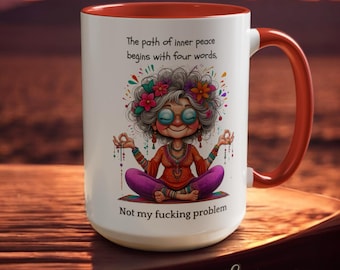 May include: A white ceramic mug with a bright orange interior and handle. The front features a colorful illustration of a person with gray hair and glasses meditating in a lotus position, surrounded by flowers. Text reads: The path of inner peace begins with four words, Not my fucking problem.