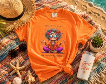 May include: An orange short-sleeved t-shirt featuring a graphic of a person with gray hair and glasses sitting in a lotus position. The text above reads "begins with four words." and below reads "Not my fucking problem."