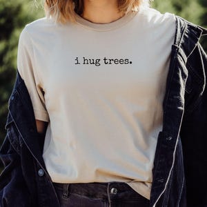 以下が含まれることがあります： 黒い文字で「i hug trees.」とプリントされたベージュのTシャツ