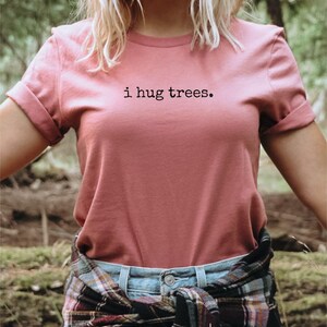 以下が含まれることがあります： 森の中で、ピンクのTシャツを着た女性。Tシャツには「i hug trees.」の文字がプリントされています。彼女は腰にチェック柄のシャツを巻いています。