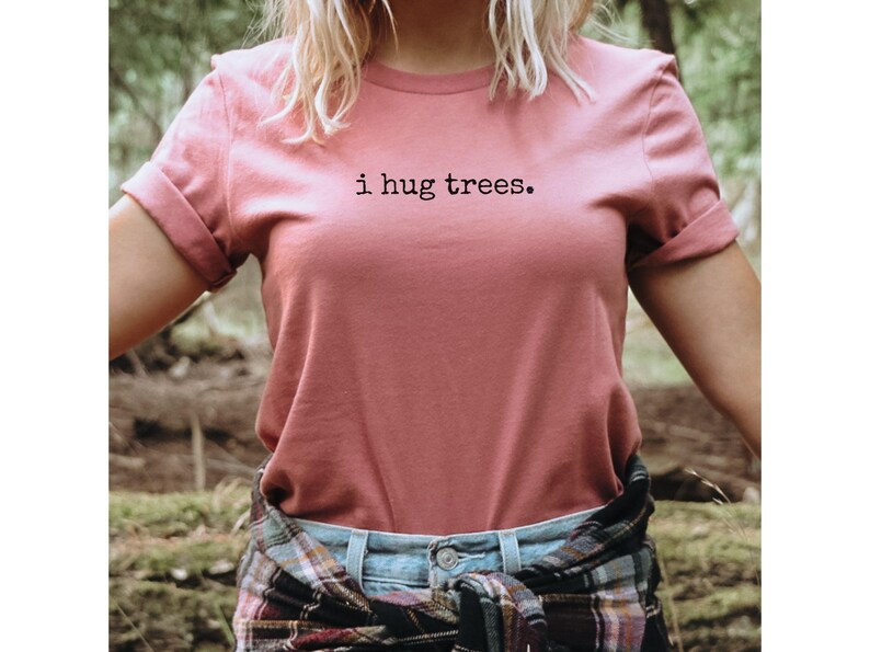 以下が含まれることがあります： 森の中で、ピンクのTシャツを着た女性。Tシャツには「i hug trees.」の文字がプリントされています。彼女は腰にチェック柄のシャツを巻いています。