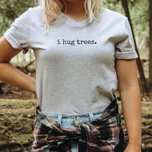 以下が含まれることがあります： VネックのグレーのTシャツで、前面に「i hug trees.」の文字がプリントされています。シャツは、腰にチェック柄のシャツを巻いた人が着ています。
