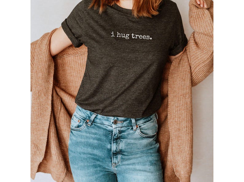 May include: A woman wearing a dark gray t-shirt with the text "i hug trees." printed in white. She is also wearing a brown cardigan and blue jeans.