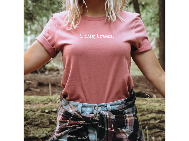 May include: A woman wearing a pink t-shirt with the text "i hug trees." printed in white. She is wearing a plaid shirt tied around her waist and is standing in a forest.