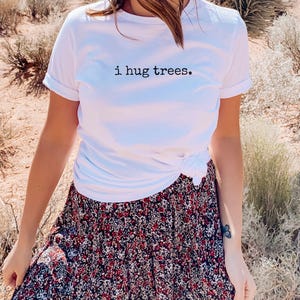 以下が含まれることがあります： 前面に「i hug trees.」という文字がプリントされた白いTシャツ。シャツはウエストで結ばれており、花柄のスカートと合わせています。