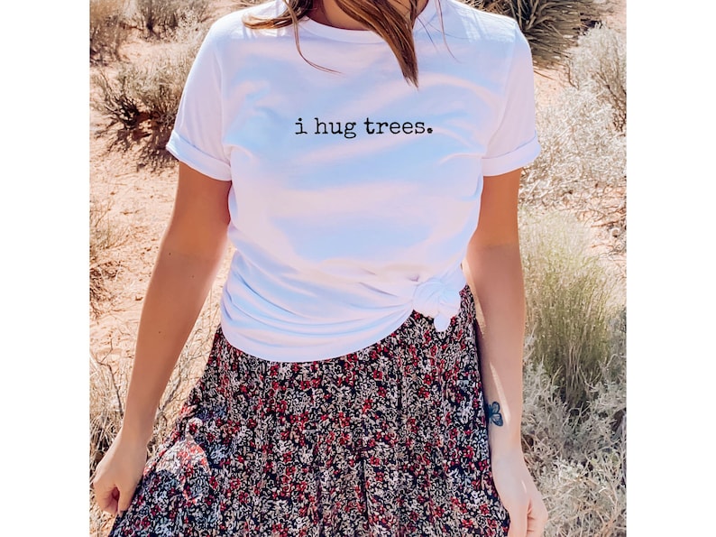 以下が含まれることがあります： 前面に「i hug trees.」という文字がプリントされた白いTシャツ。シャツはウエストで結ばれており、花柄のスカートと合わせています。