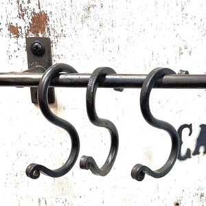 May include: A wrought iron towel rack with three S-shaped hooks. The rack is mounted on a distressed white wooden surface. The text "CARTER CONLEY" and "IRONWORKS" are stenciled on the wood.