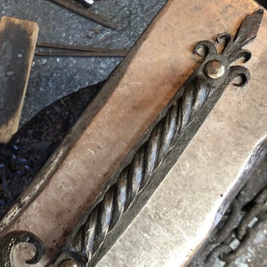 May include: A hand-forged metal door handle with a twisted design and decorative scrollwork at each end. The handle is made of dark metal and has a rustic finish.