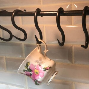 May include: A set of five black metal hooks mounted on a black metal bar. A white teacup with pink floral designs hangs from one of the hooks.