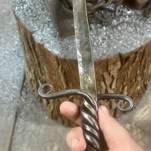 May include: Hand-forged metal sword with a twisted handle and a ring at the base. The blade is textured, and the guard features decorative swirls. The sword is held near an anvil and a wooden stump.