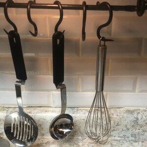 May include: Three kitchen utensils hanging on a black metal rack. The utensils include a slotted spoon, a ladle, and a whisk. The rack is mounted on a white tiled wall.