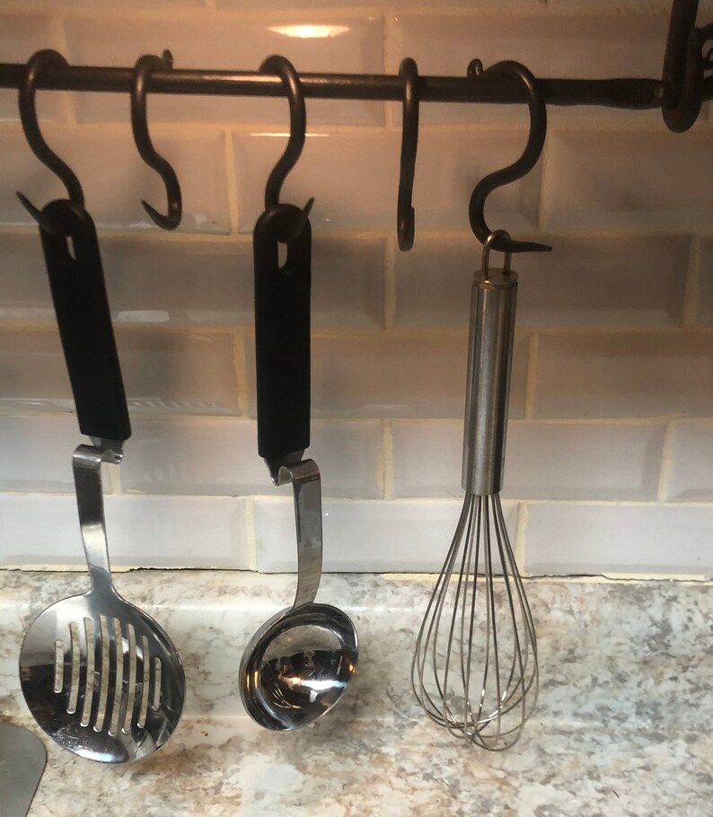 May include: Three kitchen utensils hanging on a black metal rack. The utensils include a slotted spoon, a ladle, and a whisk. The rack is mounted on a white tiled wall.