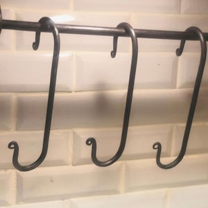 May include: Three black metal hooks hang from a metal bar against a white tiled background. The hooks are curved and have a simple design.