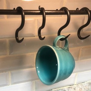 May include: A set of five black metal hooks mounted on a black metal bar, with a teal ceramic mug hanging from one of the hooks.