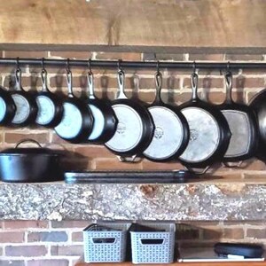 May include: A black metal rack with hooks holding a collection of cast iron cookware, including skillets, pots, and a wok. The rack is mounted on a brick wall above a wooden shelf.