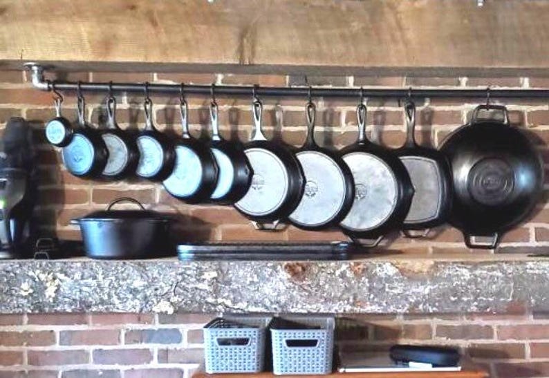 May include: A black metal rack with hooks holding a collection of cast iron cookware, including skillets, pots, and a wok. The rack is mounted on a brick wall above a wooden shelf.