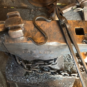 May include: A blacksmith's anvil with a metal chain wrapped around it. There is a metal hook, a hammer, and a pair of tongs on the anvil. The anvil is labeled "102 KG".