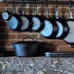 May include: A collection of cast iron cookware hanging on a black metal rack. The cookware includes a variety of sizes and shapes, including a large oval Dutch oven.