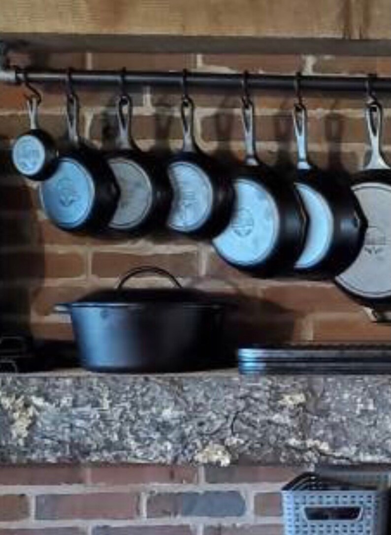 May include: A collection of cast iron cookware hanging on a black metal rack. The cookware includes a variety of sizes and shapes, including a large oval Dutch oven.
