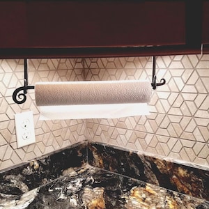 May include: A black metal paper towel holder mounted under a cabinet, with a white paper towel roll. The holder has a decorative scroll design. The holder is mounted on a beige tile backsplash with a geometric pattern.