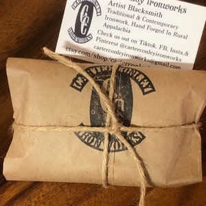 May include: A brown paper package tied with twine and a black and white logo that says "Carter Conley Ironworks". The logo is a stylized "C" and "C" with the words "Carter Conley Ironworks" wrapped around it. The package is sitting on a wooden surface.