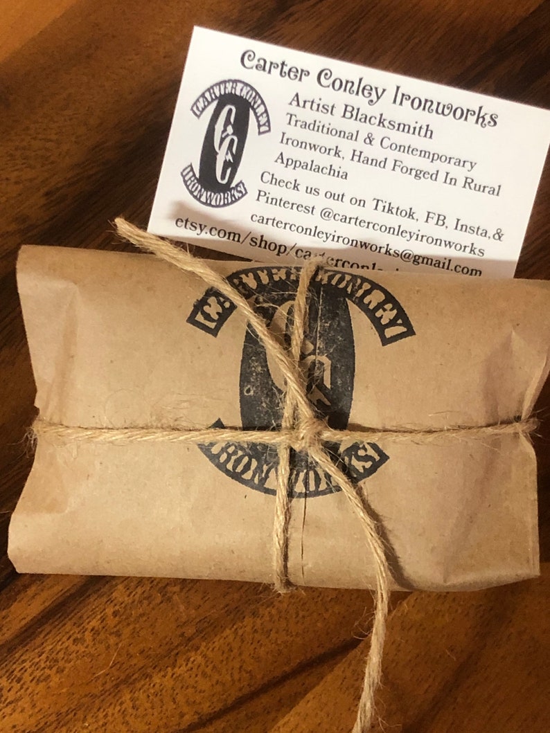 May include: A brown paper package tied with twine and a black and white logo that says "Carter Conley Ironworks". The logo is a stylized "C" and "C" with the words "Carter Conley Ironworks" wrapped around it. The package is sitting on a wooden surface.
