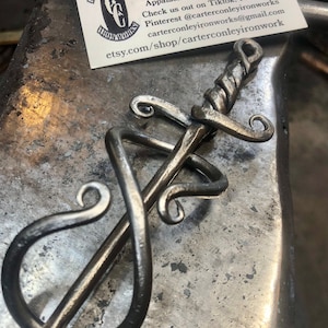 May include: A hand-forged iron sword with a twisted blade and decorative scrolls. The sword is resting on a metal surface. Carter Conley Ironworks, Artist Blacksmith, Traditional & Contemporary Ironwork, Hand Forged In Rural Appalachia. Check us out on Tiktok, FB, Insta, & Pinterest @carterconleyironworks carterconleyironworks@gmail.com etsy.com/shop/carterconleyironwork
