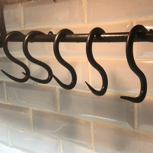 May include: A set of six black metal hooks hanging from a metal bar. The hooks are in a row and are all the same size. The hooks are attached to a white tiled wall.