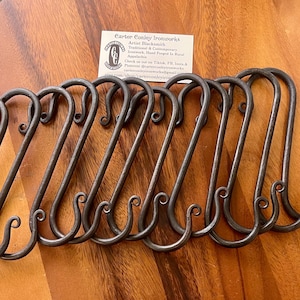 May include: Ten black metal hooks with a decorative swirl at the end of each hook. The hooks are arranged in a row on a wooden surface.
