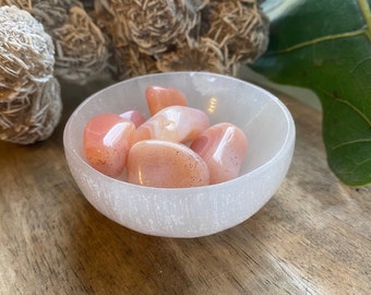 May include: A white selenite bowl filled with six carnelian tumbled stones. The stones are a light orange color with white and brown markings.