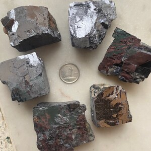 Pu&ograve; includere: Sei esemplari minerali grigio scuro di forma irregolare con lucentezza metallica sono disposti attorno a una moneta. I minerali hanno un aspetto a blocchi e angolare, con alcuni che mostrano accenni di colorazione rosso-marrone. La moneta &egrave; color argento.