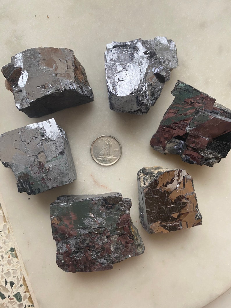Pu&ograve; includere: Sei esemplari minerali grigio scuro di forma irregolare con lucentezza metallica sono disposti attorno a una moneta. I minerali hanno un aspetto a blocchi e angolare, con alcuni che mostrano accenni di colorazione rosso-marrone. La moneta &egrave; color argento.