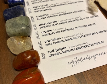 May include: A set of seven chakra stones in a pouch. The stones are amethyst, lapis lazuli, sodalite, labradorite, citrine, carnelian, and red jasper. The stones are arranged in a row from top to bottom, with the amethyst stone at the top and the red jasper stone at the bottom. The stones are all different colours and sizes. The pouch is made of a natural fabric and has a drawstring closure.