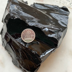 May include: A large, jagged piece of black obsidian with a glossy, reflective surface. A silver-coloured Canadian ten-cent coin from 2012 is placed on top of the stone to provide a sense of scale for the specimen's dimensions.