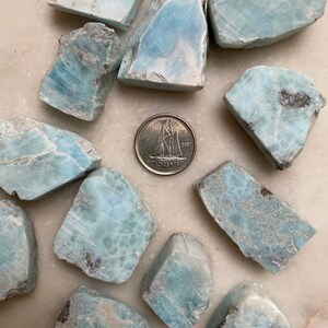 May include: A collection of 12 blue and white polished blue lace agate stones. The stones are various shapes and sizes, and some have a cloudy appearance. A Canadian dime is included for scale.