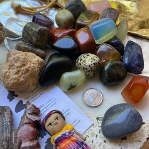 Peut inclure: Une collection de pierres précieuses polies de différentes couleurs, dont le vert, le marron et le bleu, ainsi qu'une petite poupée, une figurine d'écureuil sculptée et une pièce de monnaie. Un sac doré à cordon est visible en arrière-plan. Une carte avec du texte est également présente.