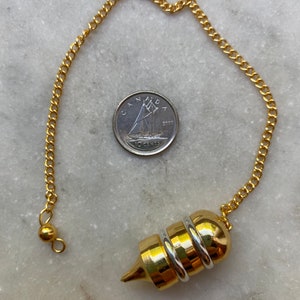 May include: A gold-colored pendulum with a pointed tip and multiple rings around the center. The pendulum is attached to a gold chain with a small ball at the end.