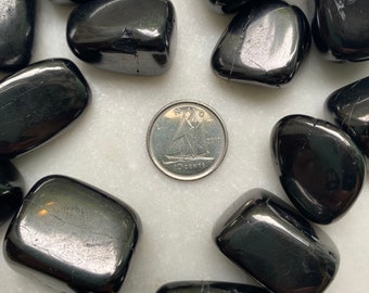 May include: A collection of black obsidian tumbled stones, some with visible striations, arranged in a heart shape on a white surface. A Canadian 10-cent coin is placed in the center for scale.