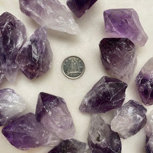 May include: A collection of amethyst crystals in various sizes and shapes. The crystals are a deep purple color with some lighter areas. The crystals are arranged in a circle around a Canadian 10 cent coin.