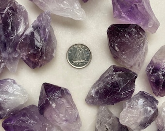 May include: A collection of amethyst crystals in various sizes and shapes. The crystals are a deep purple color with some lighter areas. The crystals are arranged in a circle around a Canadian 10 cent coin.