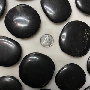 Peut inclure: Une collection de pierres d'obsidienne noires lisses, certaines avec un léger éclat, disposées sur une surface blanche. Une pièce de monnaie canadienne est placée au centre pour l'échelle.