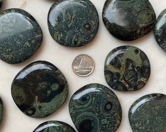 May include: A collection of polished green and black ocean jasper stones, each with a unique swirling pattern. The stones are smooth and oval-shaped, and they are arranged on a white surface.