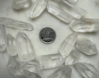May include: A collection of clear quartz crystal points of varying sizes and shapes. The crystals are scattered on a white surface, with a Canadian 10-cent coin in the center for size reference. The crystals have a translucent appearance.