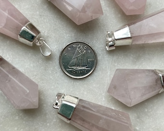 May include: A collection of rose quartz crystal pendants with silver-colored metal caps. The pendants are a soft pink color and have a faceted, elongated shape. A Canadian 10-cent coin is included for size reference. The pendants are arranged on a light-colored surface.