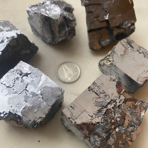 Pu&ograve; includere: Sei campioni di minerale grigio scuro, dall'aspetto metallico, con forme angolari, sono disposti attorno a una moneta. I minerali hanno una superficie ruvida e strutturata e riflettono la luce. La moneta &egrave; d'argento e fornisce un riferimento dimensionale.