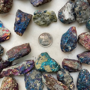 May include: A collection of iridescent pyrite stones, also known as fool's gold, in various sizes and shapes. The stones have a metallic sheen with shades of blue, purple, and gold. A Canadian 10-cent coin is included for scale.