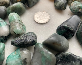 May include: A collection of tumbled emerald stones with black inclusions. The stones are various shades of green and gray, and some have a translucent quality. A Canadian dime is included for scale.